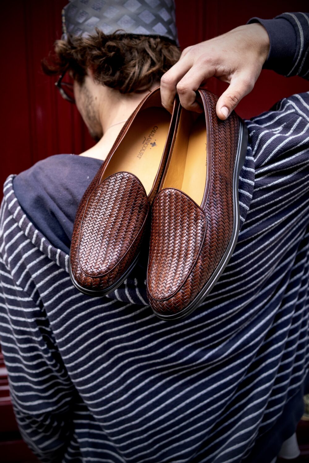 A person holds a pair of brown woven loafers while wearing a striped hoodie.