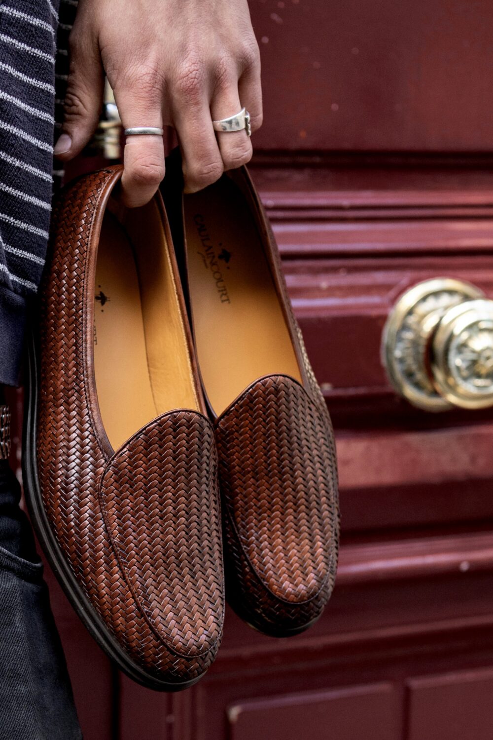 A person holds a pair of brown woven slip-on shoes with a tan interior.