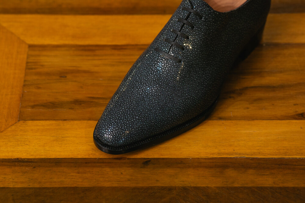 A close-up of a dark gray, textured dress shoe with a pointed toe and laces.