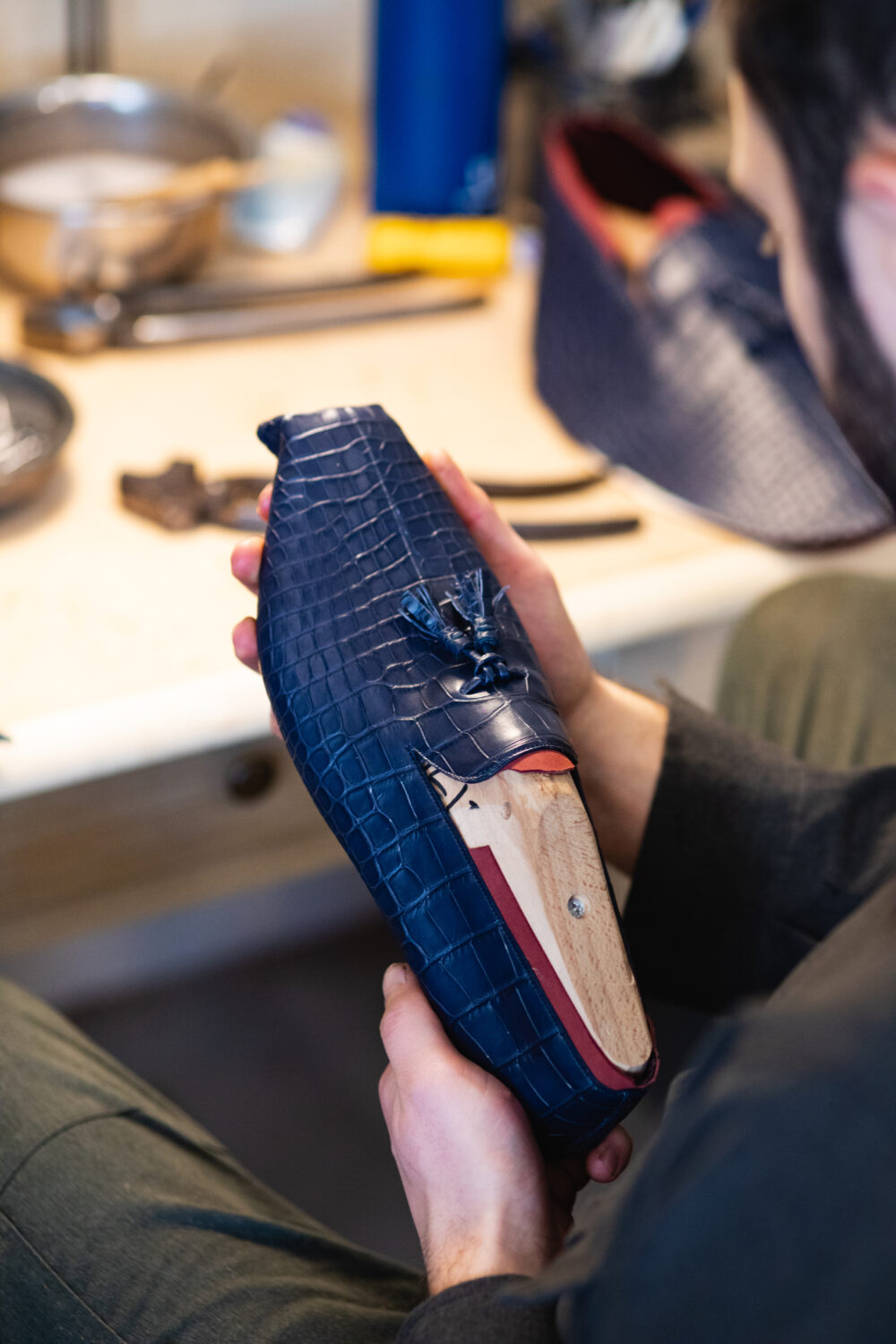 A person holds a navy blue crocodile-patterned shoe upper with a wooden shoe last inside it.