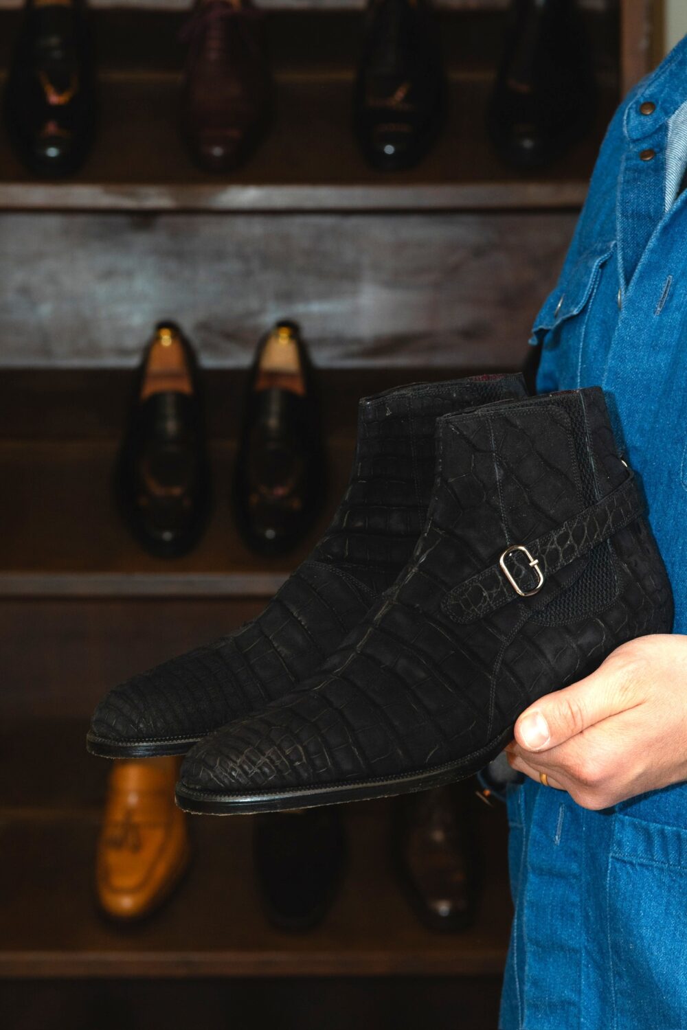 A person holds a pair of black crocodile-textured ankle boots with a buckle.