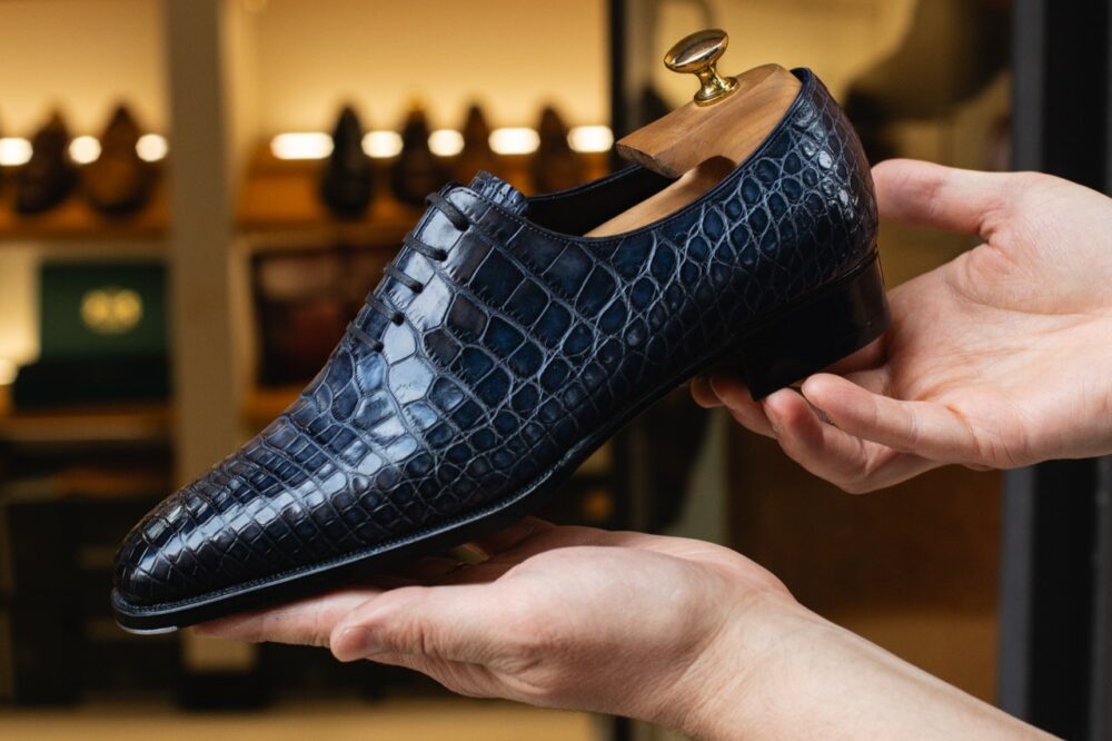 A dark blue crocodile leather shoe is being held by two hands.