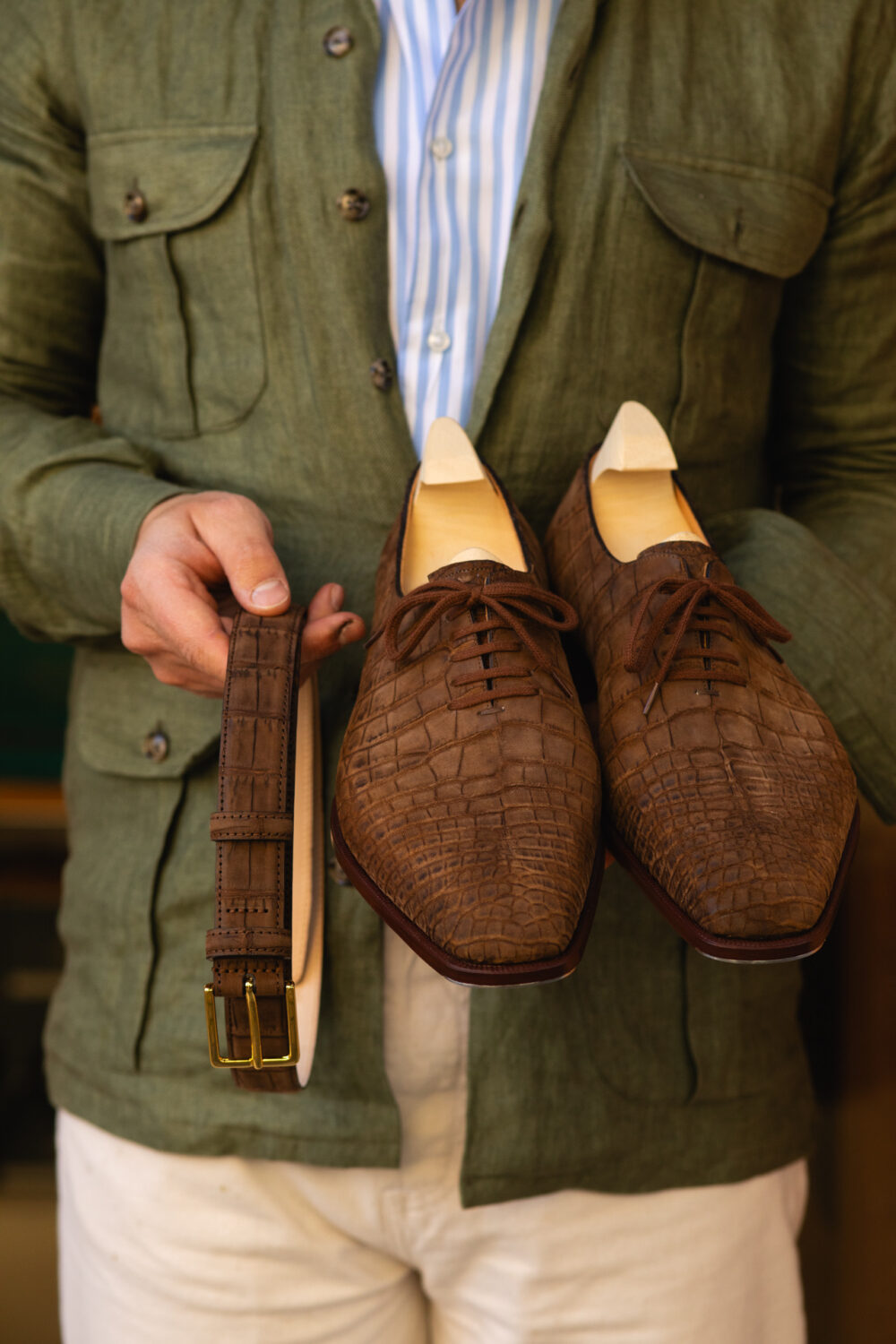 A person holds a pair of brown crocodile leather shoes and a matching brown belt.
