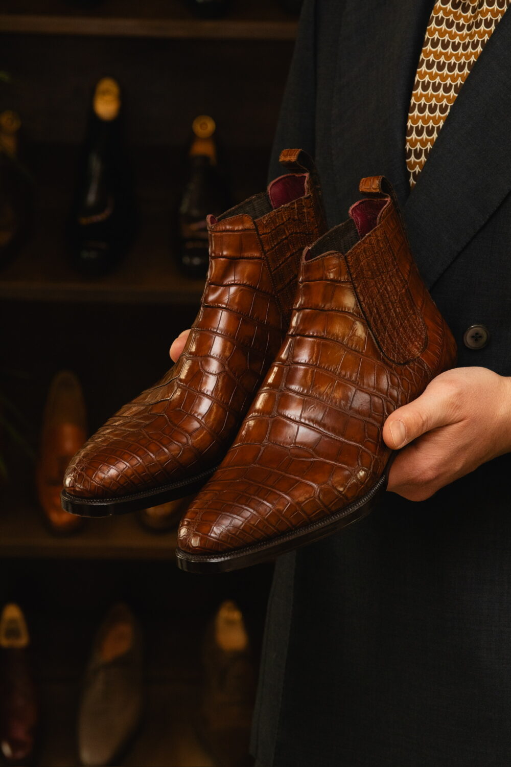 A person holds a pair of brown crocodile leather chelsea boots.