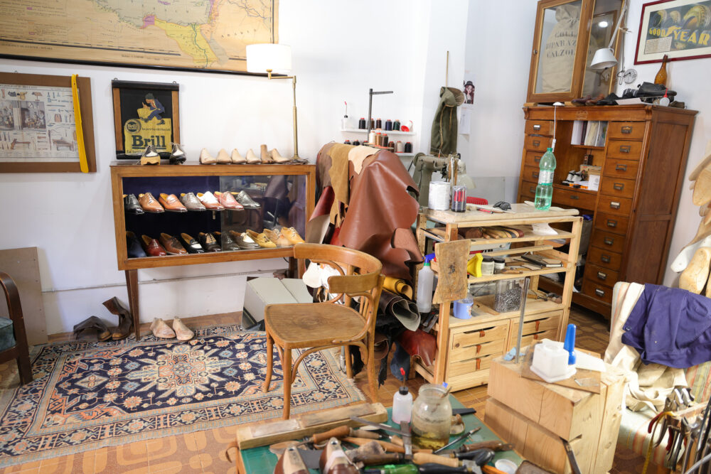 A cluttered shoemaker's workshop featuring a display case of various leather shoes, wooden tools, and rolls of leather.