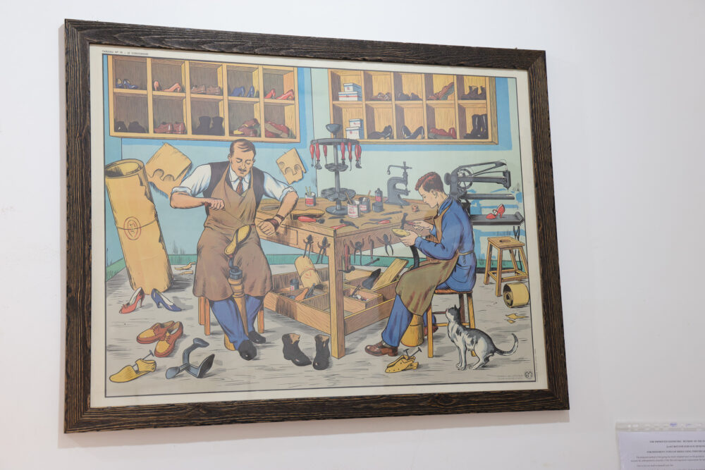 Illustration of two shoemakers working at a wooden table, one carving leather and the other reading, with various shoes scattered around and a cat nearby.