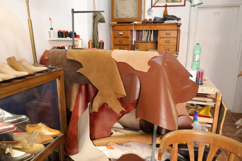 A selection of leather pieces in various shades of brown and tan is displayed on a worktable alongside wooden shoe forms.
