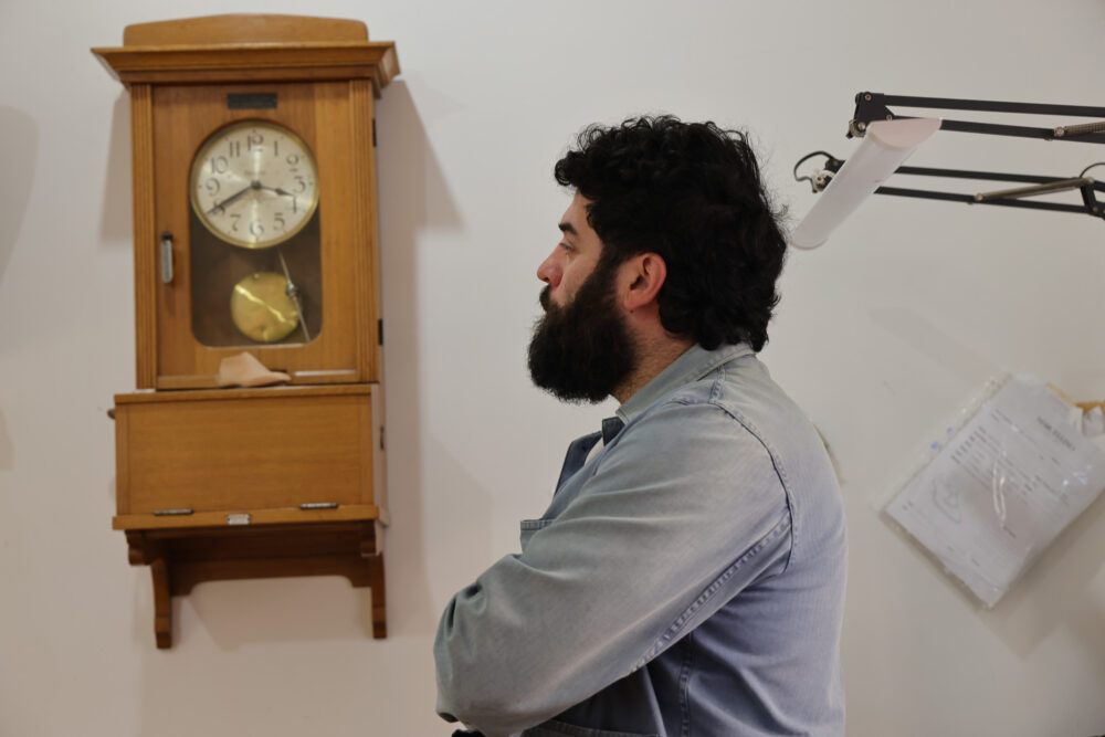 A man with a beard and curly hair is sitting with his arms crossed, next to a wooden wall clock.