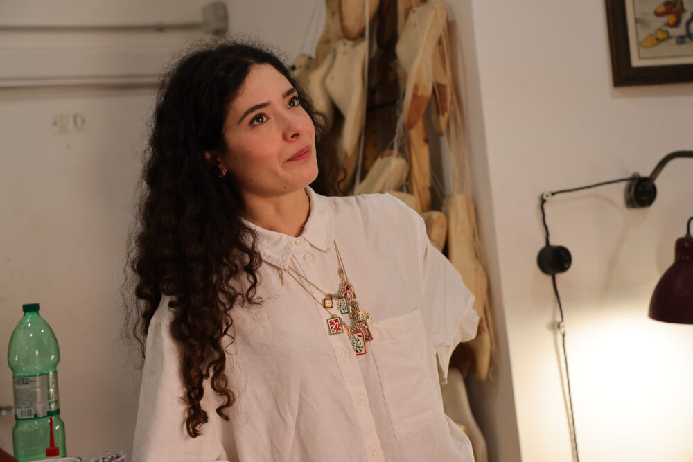 A woman with curly hair wears a white shirt and multiple colorful necklaces while looking thoughtfully to the side.