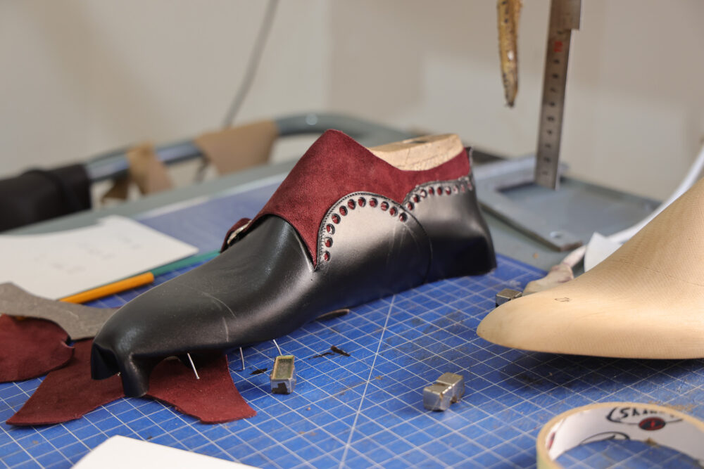 A black and burgundy shoe mold with decorative studs is placed on a blue cutting mat, alongside leather scraps and tools.