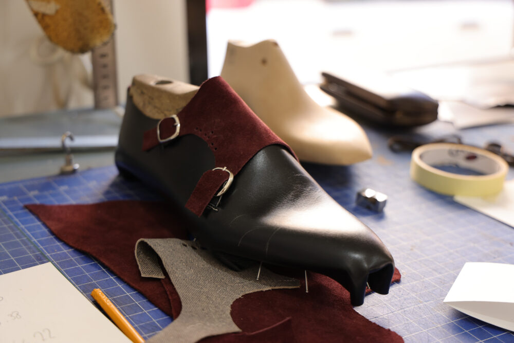 A black leather shoe with burgundy suede accents is shown on a work surface surrounded by various materials and tools.
