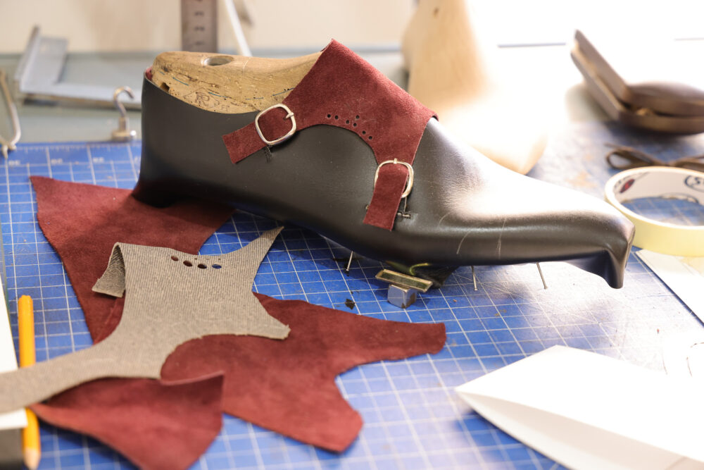 A black shoe mold with red suede patches and leather pieces is surrounded by crafting materials on a blue cutting mat.
