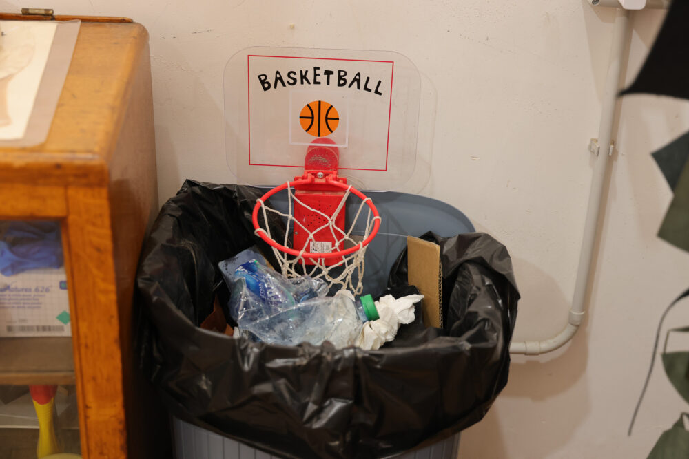 A trash bin with a red basketball hoop attached, filled with plastic bottles and crumpled paper.