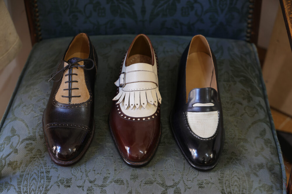 Three pairs of handcrafted shoes: a black lace-up with a beige toe, a brown slip-on with a white fringed strap, and a black loafer with a textured white panel.