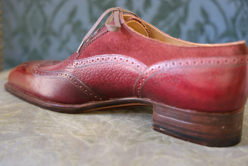 A close-up of a red leather dress shoe with decorative perforations and a brown wooden heel.