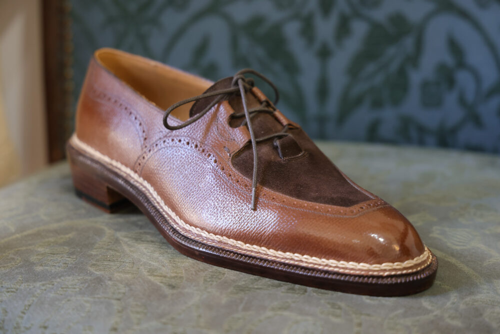 A brown leather shoe with a contrasting dark brown suede toe and decorative stitching.