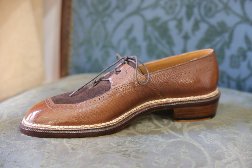 A polished brown leather shoe with a suede toe cap and decorative brogue detailing.