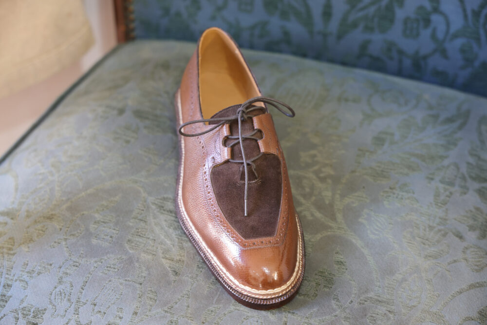 A brown leather shoe with a suede accent and laces sits on a textured green surface.