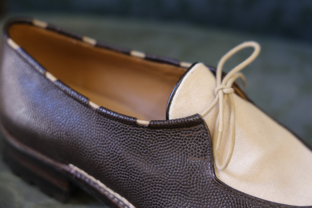 A close-up of a brown leather shoe with a cream-colored canvas toe and a decorative lace.