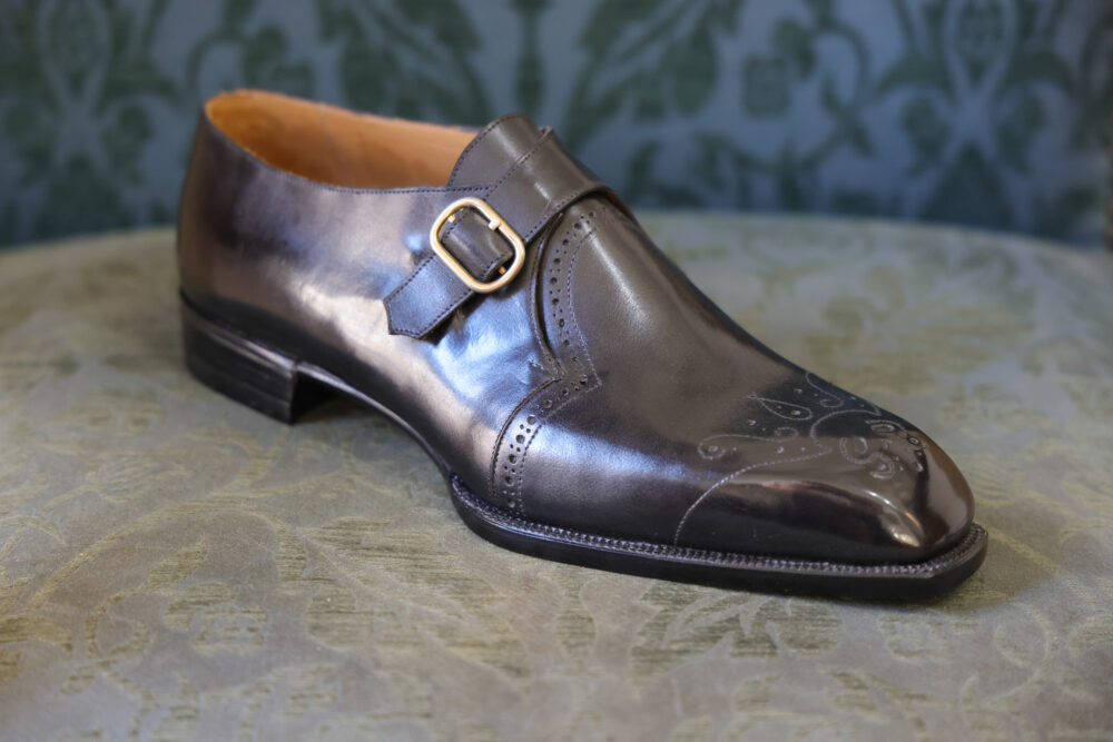 A polished dark leather shoe with a decorative buckle and intricate detailing on the toe.