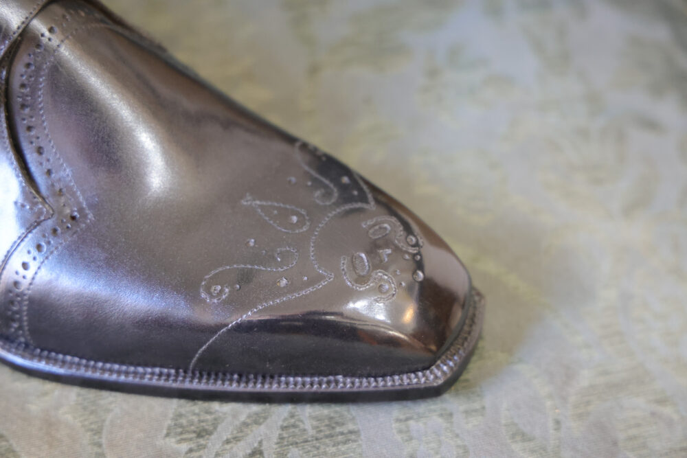 A close-up of a polished, metallic gray leather shoe featuring intricate decorative stitching on the toe.