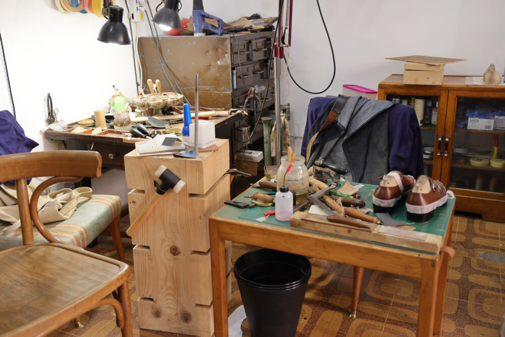 A wooden table cluttered with shoemaking tools, leather shoes, and a mallet sits beside a chair in a workshop.