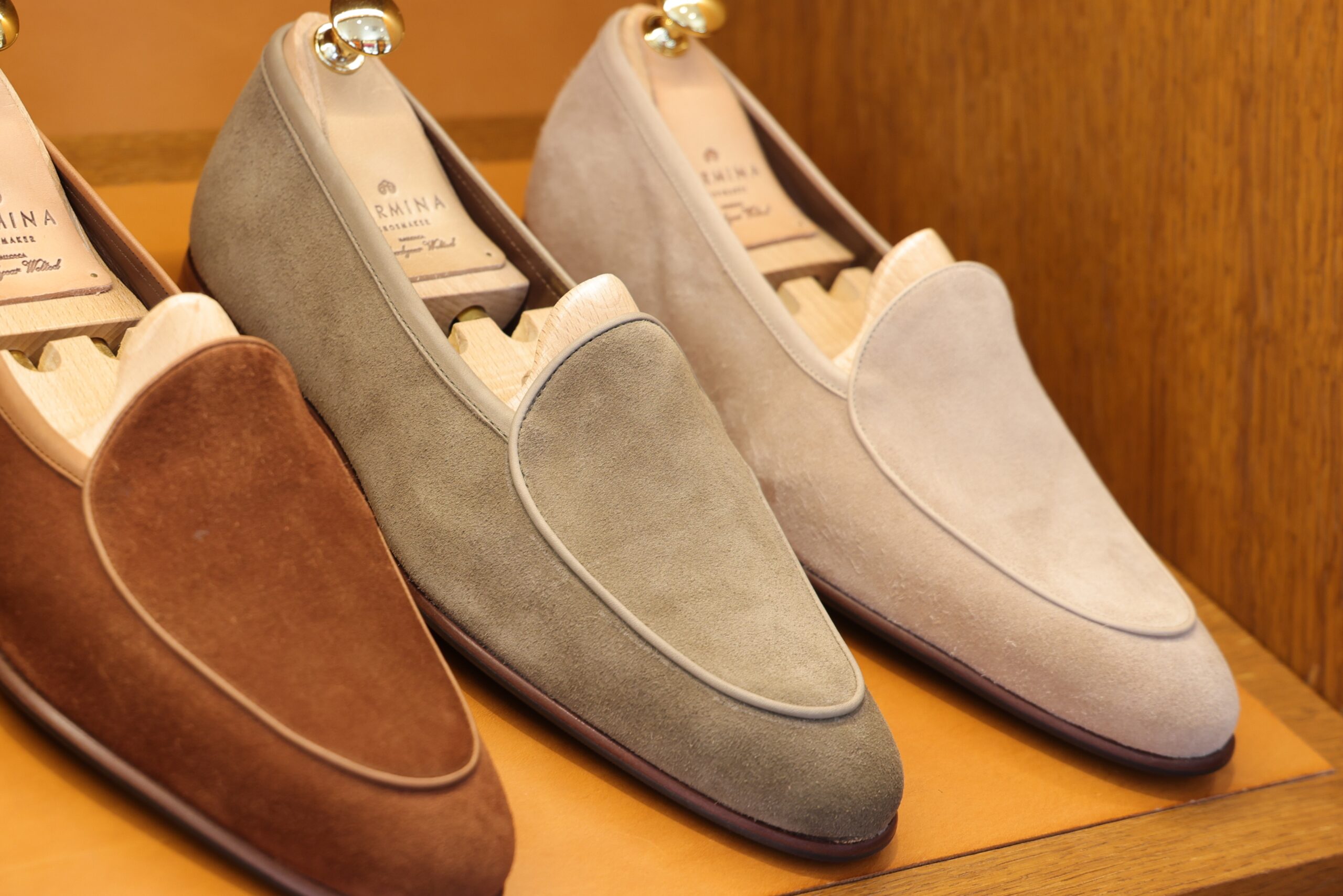 Three pairs of suede loafers in brown, green, and beige, displayed with wooden shoe trees inside.