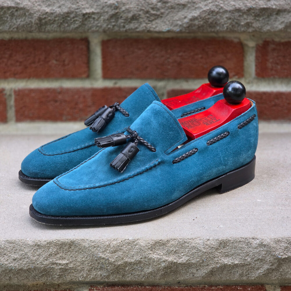 Turquoise suede loafers with black tassels and red insoles.