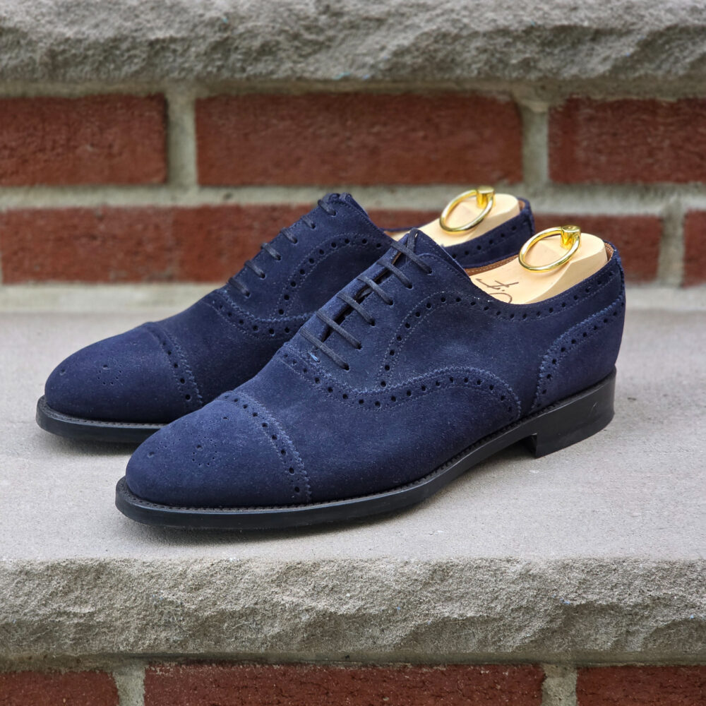 A pair of navy blue suede shoes with brogue detailing and gold shoe trees.