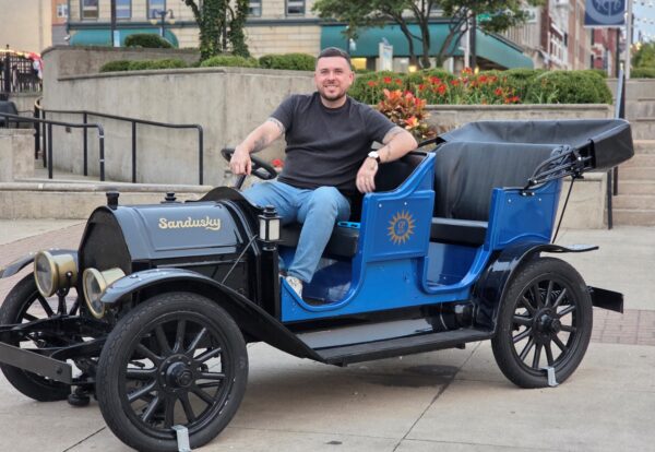 A person sits in a vintage black and blue car with the word "Sandusky" displayed on the front.