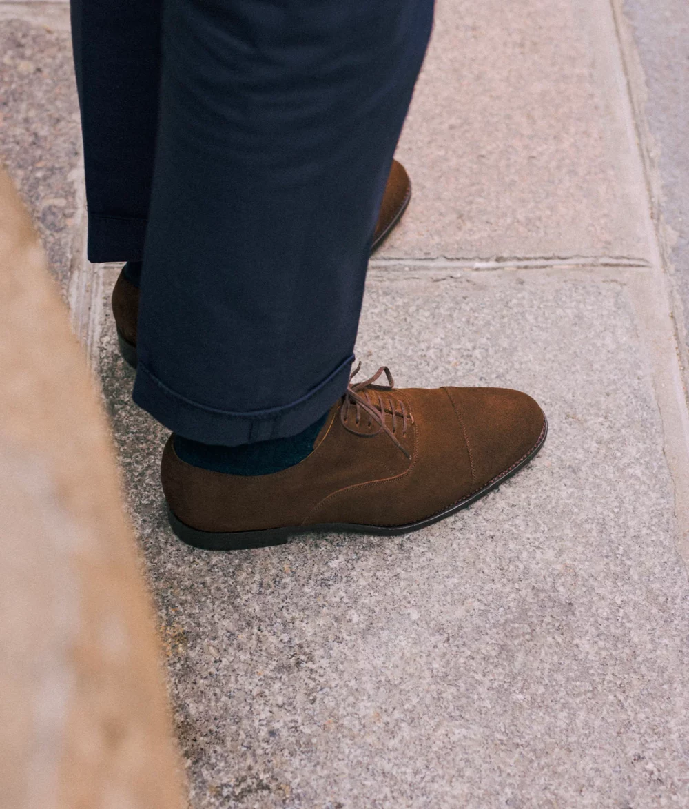 A close-up of a brown suede shoe paired with navy trousers, featured in the Septieme Largeur Summer Sale 2026.