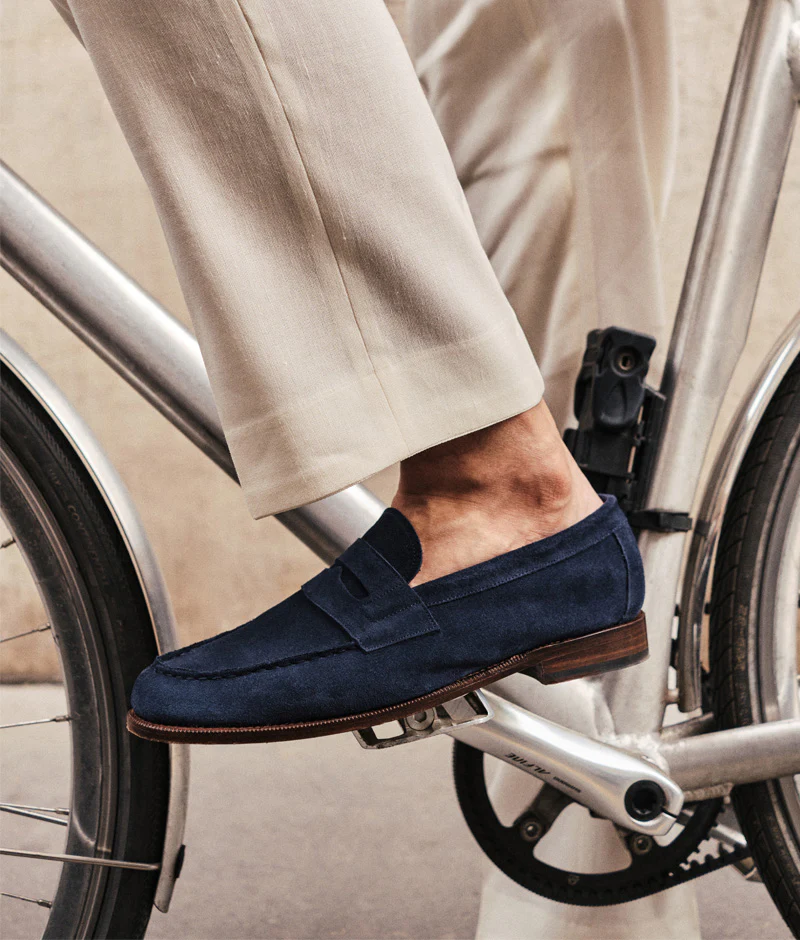 A close-up of a foot wearing navy suede loafers while resting on a bicycle pedal, featured in the Septieme Largeur Summer Sale 2026.