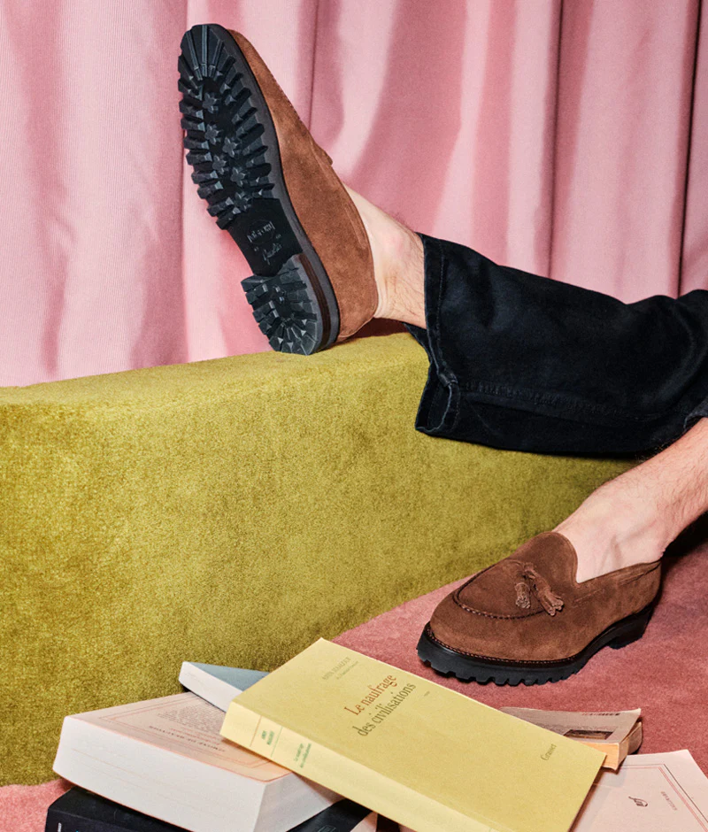 A foot wearing a brown suede loafer with a textured black sole rests on a green cushion, featured in the Septieme Largeur Summer Sale 2026.