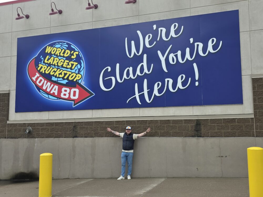 A person stands with arms outstretched in front of a large blue sign that reads "World's Largest Truckstop Iowa 80" and "We're Glad You're Here!" in yellow and white lettering.