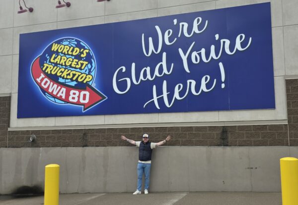A person stands with arms outstretched in front of a large blue sign that reads "World's Largest Truckstop Iowa 80" and "We're Glad You're Here!" in yellow and white lettering.