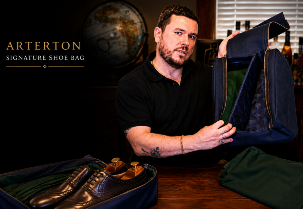 A man in a black shirt holds an open navy blue shoe bag, showcasing its green interior and a pair of brown dress shoes inside.