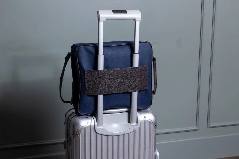 A navy blue Arterton Signature Shoe Bag is attached to the handle of a silver suitcase.