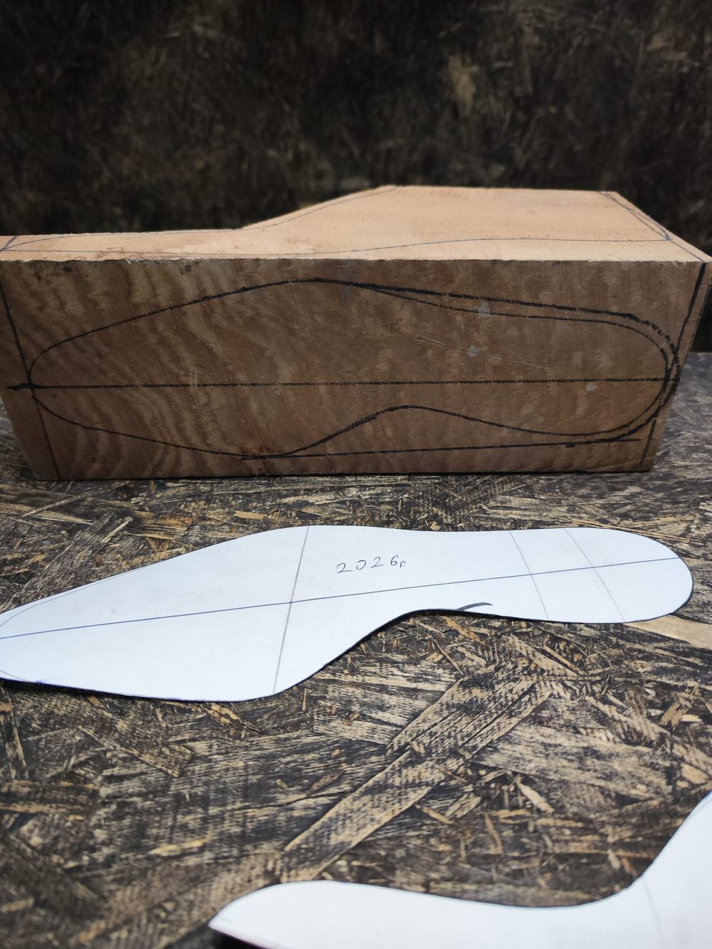 A wooden shoe last with a marked outline and two paper templates labeled "2026" in a workshop setting.