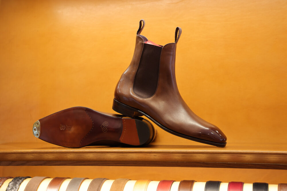 A pair of polished brown leather Chelsea boots with elastic side panels, displayed on a wooden shelf.