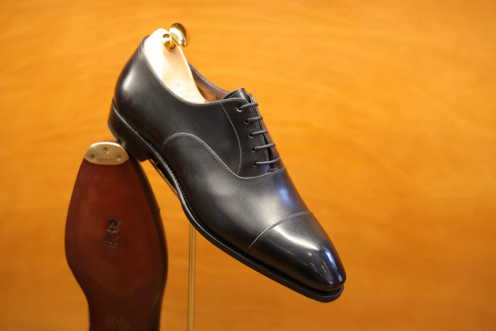 A polished black leather dress shoe displayed on a stand, showcasing its elegant design and detailed stitching.