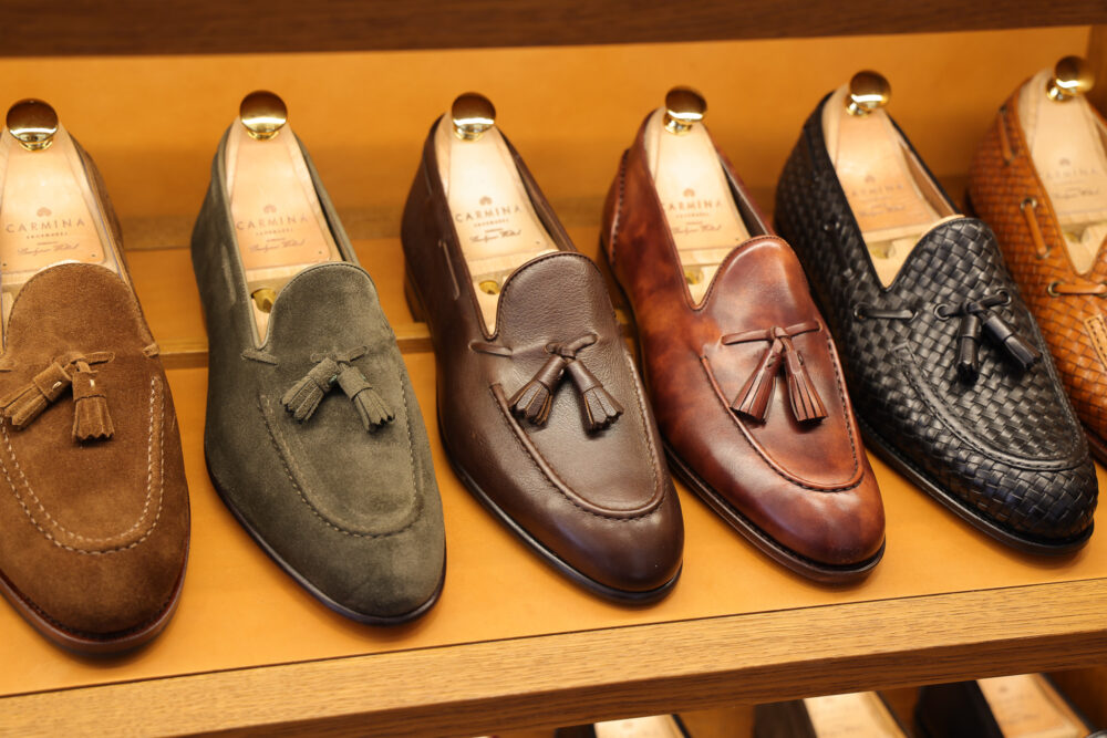 A display of five pairs of Carmina Shoemaker loafers in various colors, including brown suede, green suede, dark brown leather, tan leather, and black woven leather.