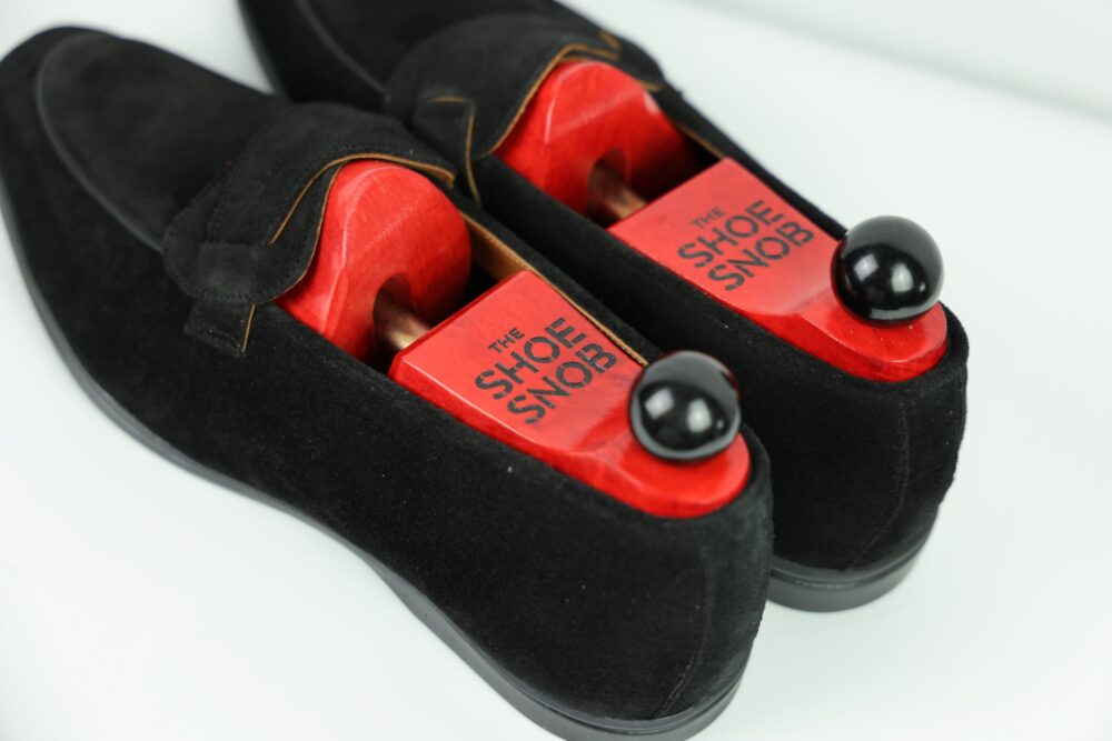 A pair of black suede shoes featuring red wooden shoe trees with a black knob labeled "THE SHOE SNOB."