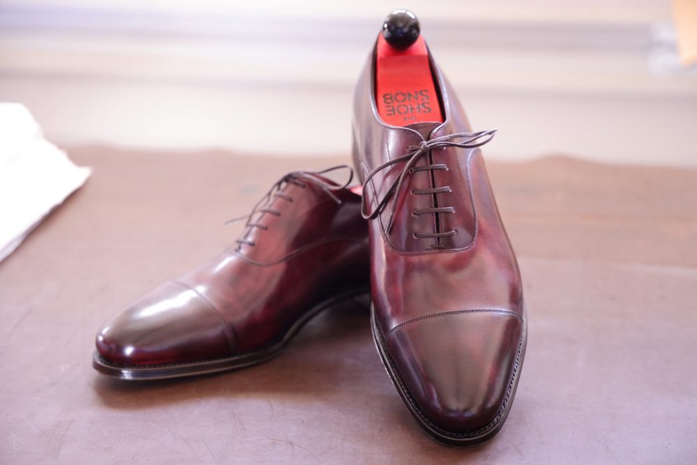 A pair of burgundy leather dress shoes featuring a cap toe and brown laces, with "Shoe Snob" visible on the insoles, alongside The Shoe Snob's Lightweight Shoe Trees.