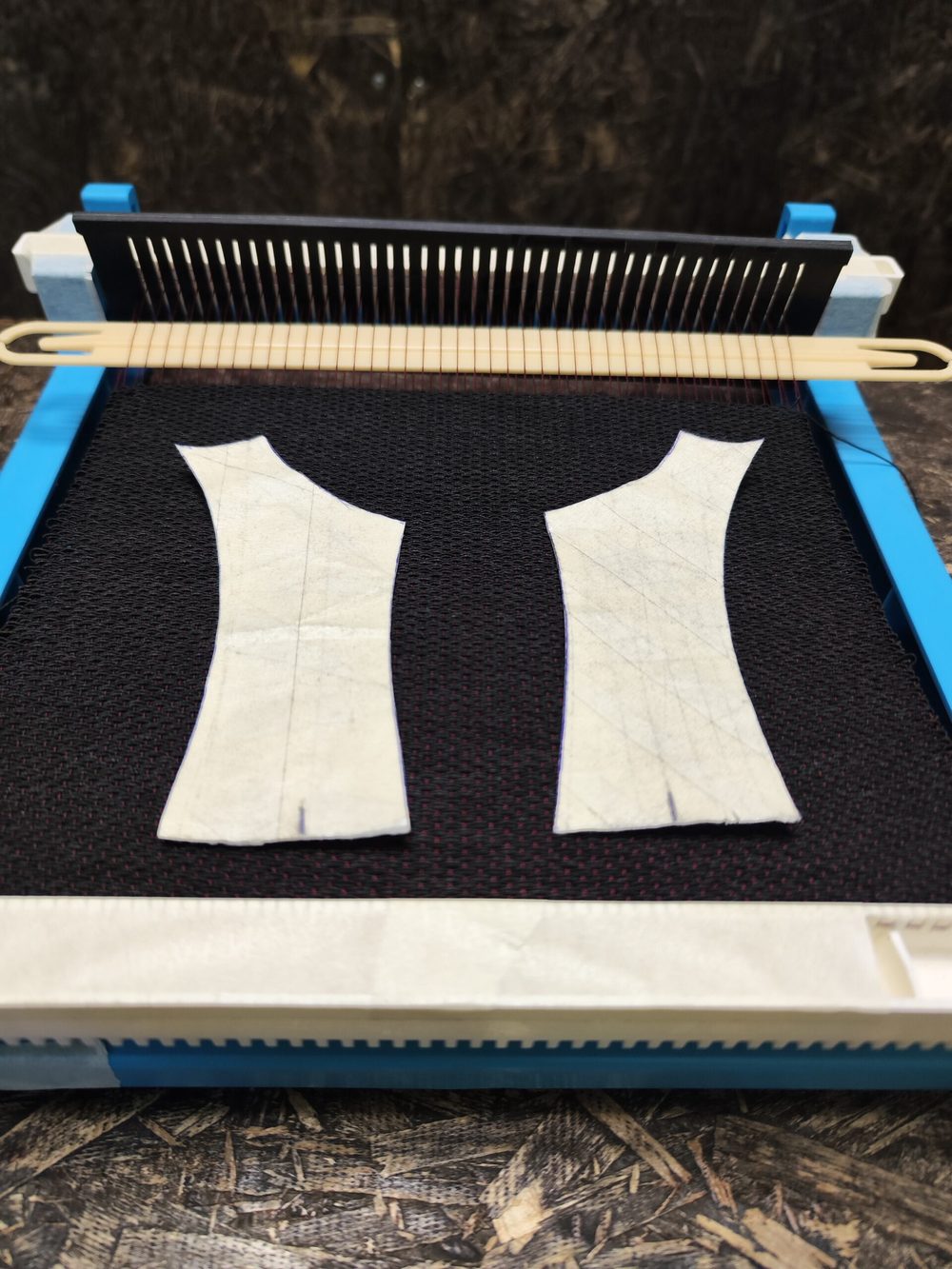 A blue shoemaking tool with two white leather patterns and a wooden needle placed on a black mesh surface.
