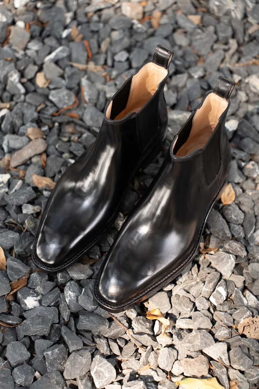 A pair of shiny black cordovan ankle boots with a pointed toe and pull-tabs.
