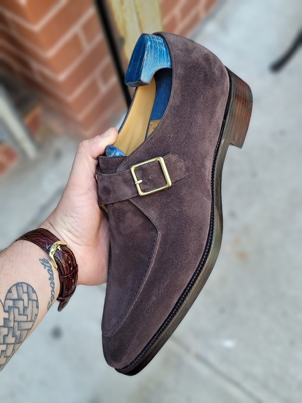 A hand holds a dark brown suede shoe with a gold buckle and a blue heel.