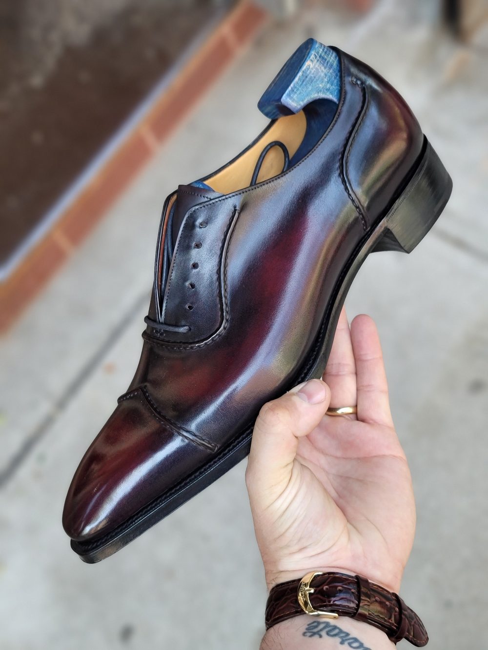 A hand holds a polished burgundy leather shoe with a blue heel and decorative stitching.