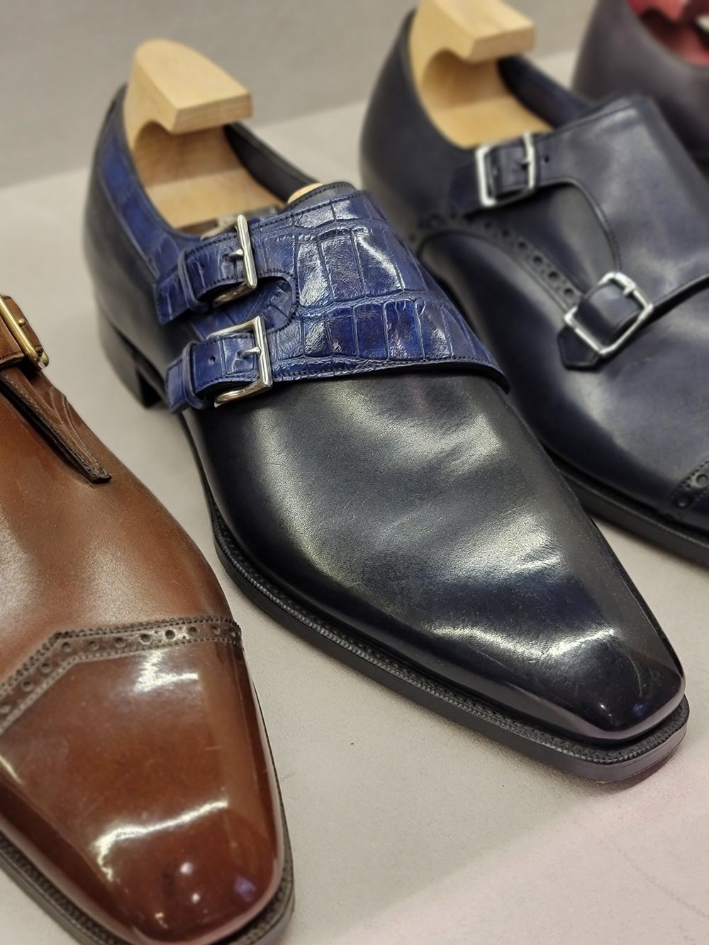 Three pairs of dress shoes are displayed, featuring a brown leather shoe with brogue detailing, a black shoe with a shiny finish and blue crocodile leather straps, and a dark blue shoe with silver buckles.