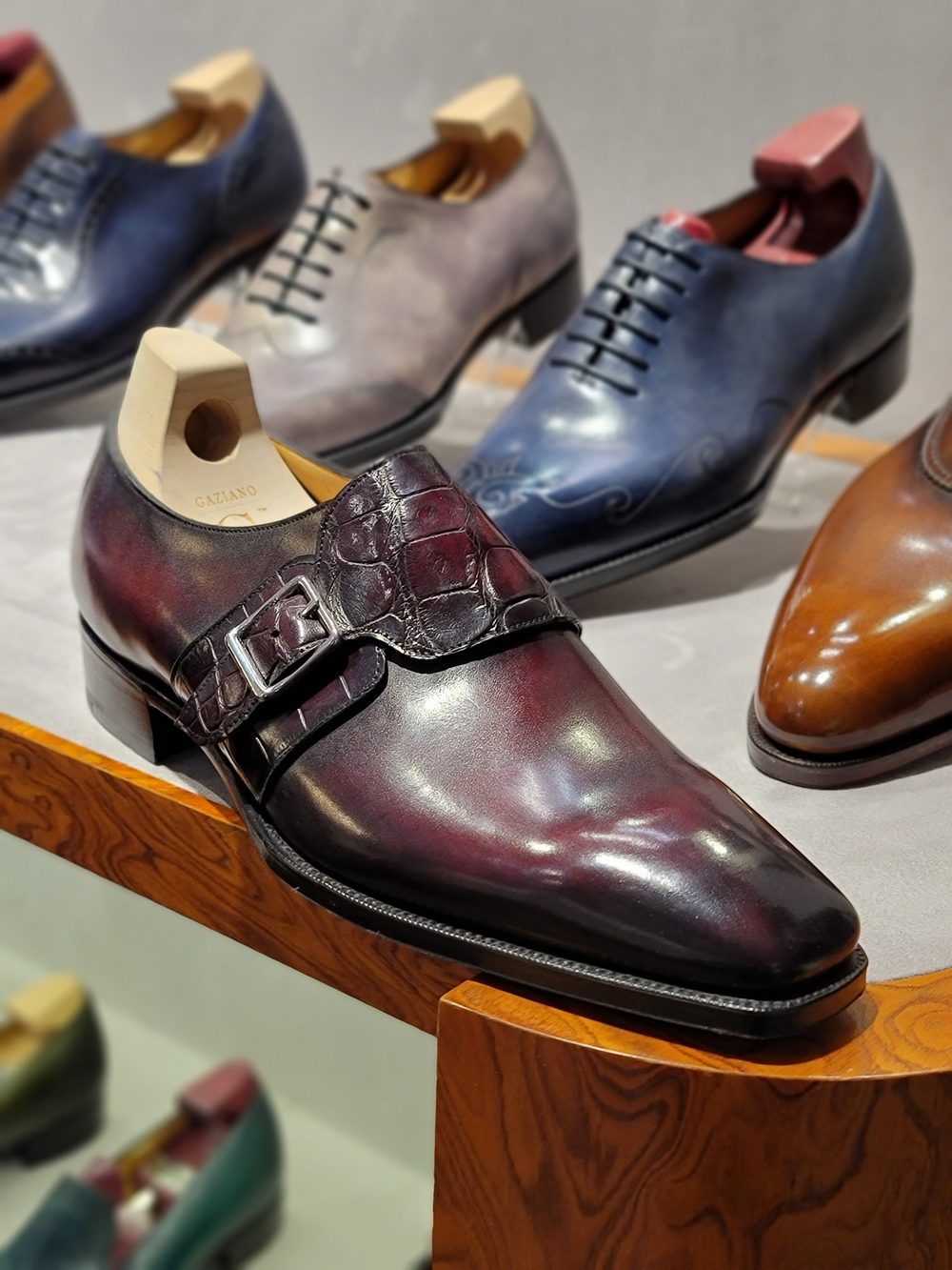 A burgundy leather shoe with a crocodile leather strap and buckle, displayed among other shoes.