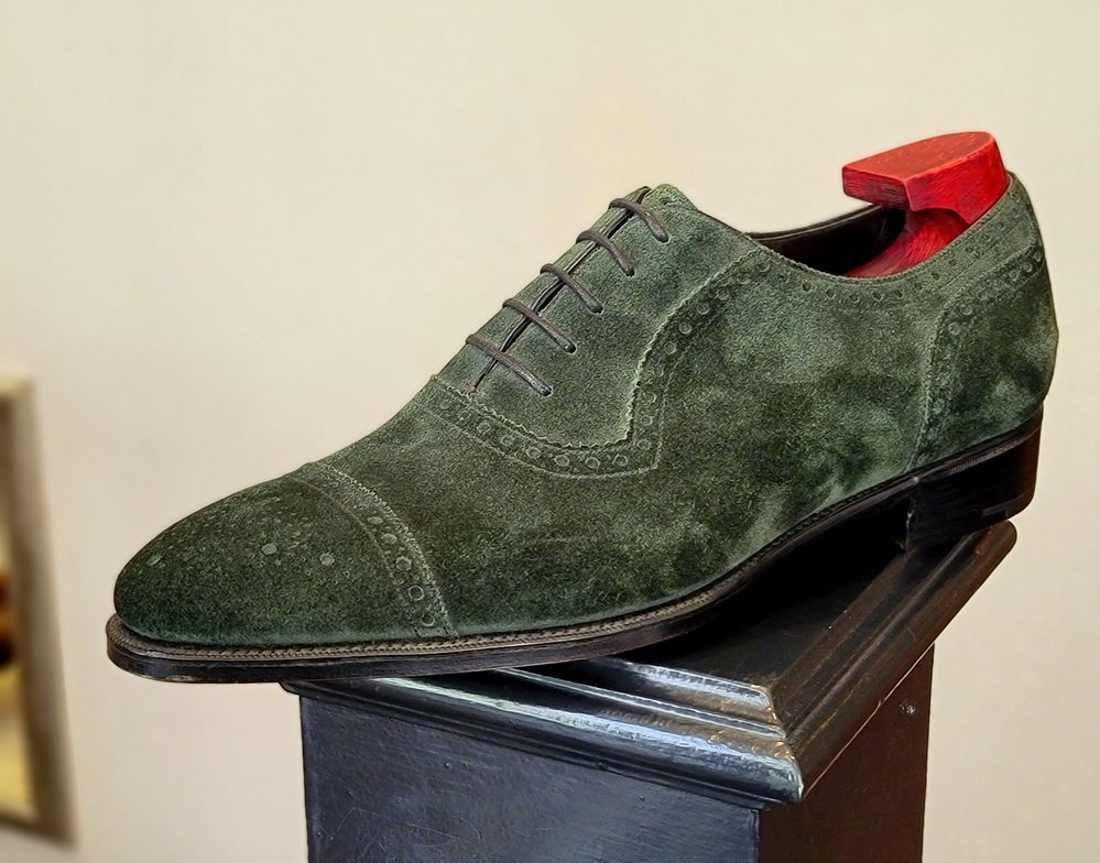 A green suede shoe with brogue detailing and a red wooden heel.